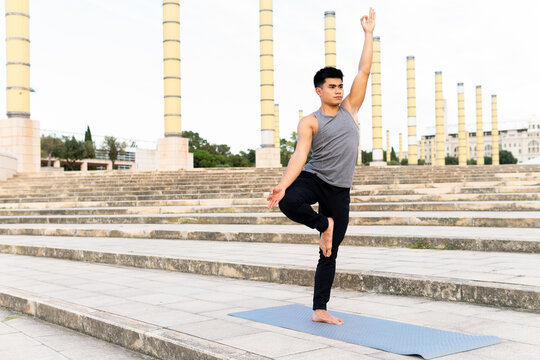 Young concentrated Asian male standing in Vrksasana with mudra gesture and doing yoga on mat on stairs in city park