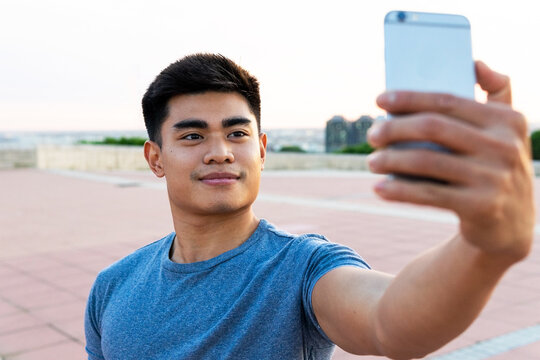 Handsome Asian male standing in city while taking photo on selfie camera of cellphone and smiling