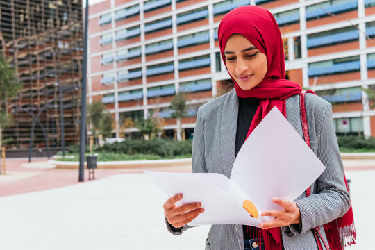 Arab Female Student In Traditional Headscarf Sitting In Street And Reading Documents For Homework