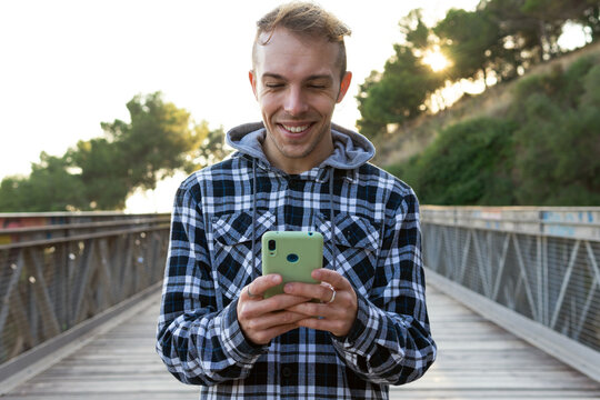 Positive young male traveler in checkered shirt checking data on smartphone while standing on wooden footbridge during summer journey in countryside