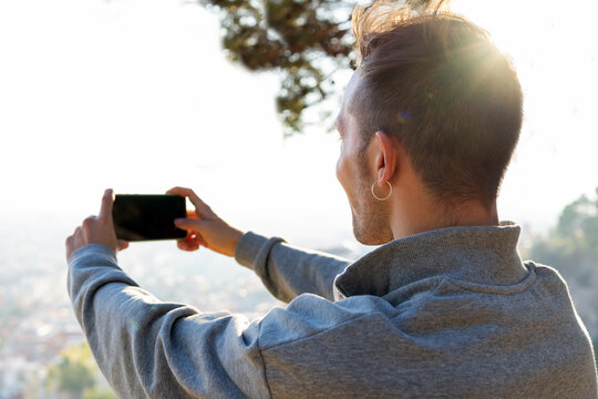 Side view of happy hipster male traveler taking pictures with mobile phone while standing on green hill against blurred background in sunny day