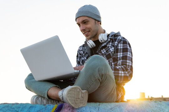 Low angle of happy young hipster male blogger in casual outfit with headphones on neck sitting on stone border and browsing laptop at sunset time