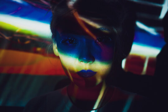 Young Asian female with bright creative makeup looking at camera while standing in dark room illuminated with colorful projector