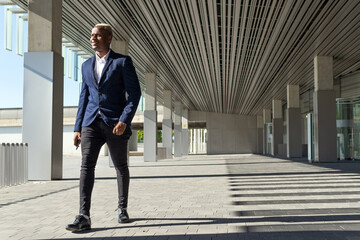 Cheerful black male entrepreneur in stylish formal outfit walking along sunny street and looking away