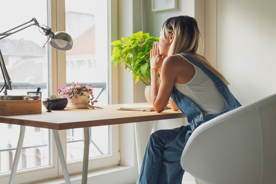 Side view of sad female leaning on hands and sitting at table at home while contemplating and looking away in frustration