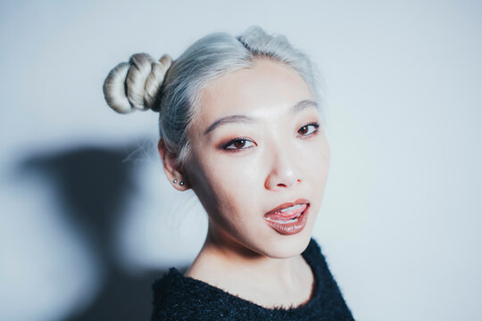 Asian teen hipster female with dyed hair looking at camera while standing under illumination against white wall with shadow
