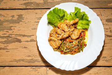 Vegetables with chicken in a pan-Asian sauce with sesame seeds on green salad leaves on a wooden table in a plate.