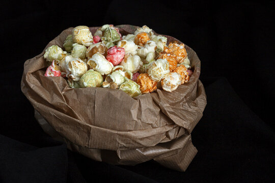Heap of varicolored sweet popcorn in paper bag placed with yellow corn seeds on black background in studio