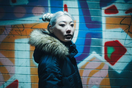 Side view of asian teen hipster female with dyed hair wearing warm jacket looking at camera while standing under illumination against colorful wall with graffiti