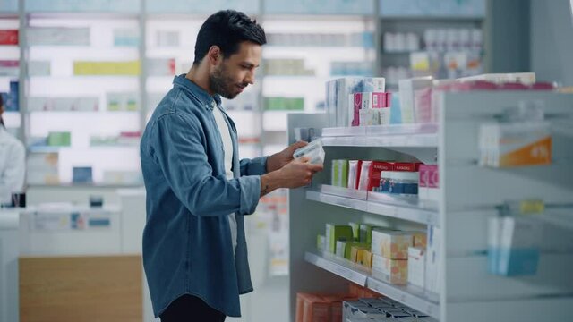 Pharmacy Drugstore: Portrait of Handsome Young Latin Man Searching to Purchase Best Medicine, Chooses between Two Packages of Drugs, Vitamins. Shelves full of Health Care, Wellness, Sport Supplements