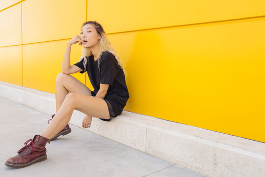 Full body side view of Asian teen female hipster in casual wear and boots sitting near yellow wall on city street