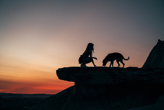 Side view of silhouette of unrecognizable female sitting on hill with dog on background of sundown in Bardenas Reales