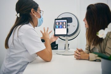 Side view of cosmetologist and customer sitting at table with facial skin diagnostics equipment while looking at tablet and discussing results of analysis in beauty clinic