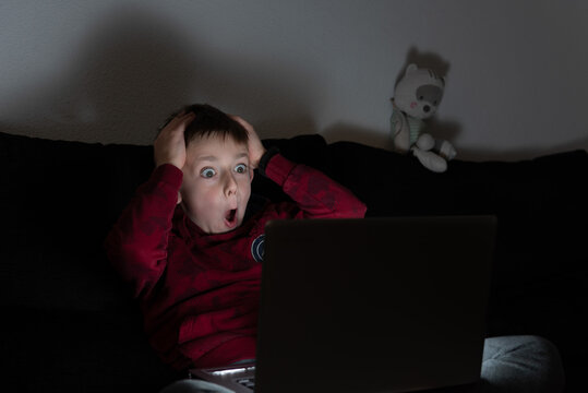 Terrified boy sitting on sofa in dark room and watching horror film on laptop