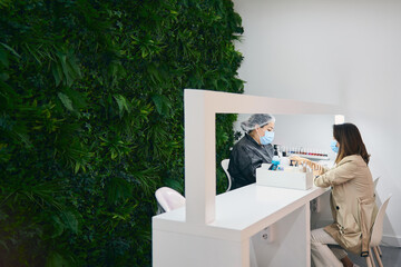 Side view of cheerful manicure master using nail file while sitting at table with female client in modern beauty salon during coronavirus pandemic