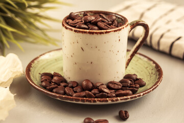 Ceramic coffee cup filled with roasted dark brown coffee beans, green grass  on light  background. Taste of coffee.  Sunny morning. Side view