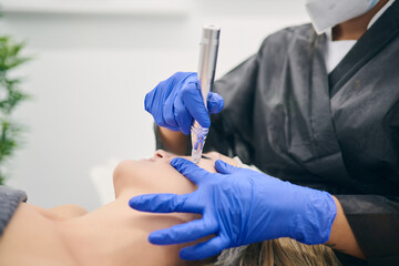 Cropped unrecognizable cosmetologist in uniform using derma pen with micro needles and cleaning face of client in beauty clinic
