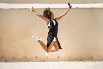 Side view of delighted African American female taking selfie on smartphone in moment of jumping above ground with outstretched arms