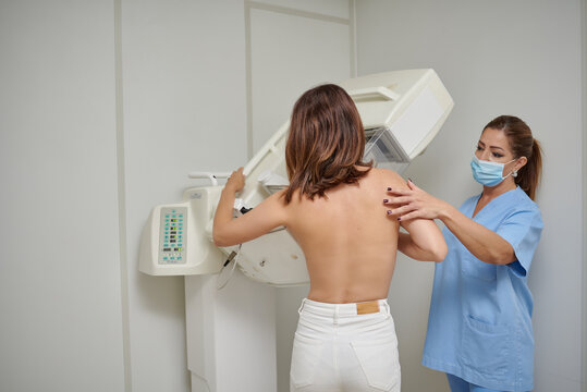 Female medic in masks and uniform preparing woman with naked torso for chest radiology while standing in medical room in clinic