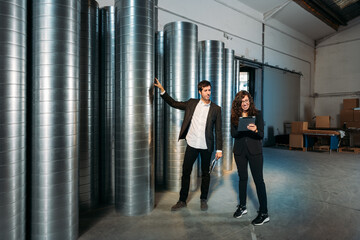 Busy male and female colleagues in formal clothes using tablet together while working in storehouse with metal barrels