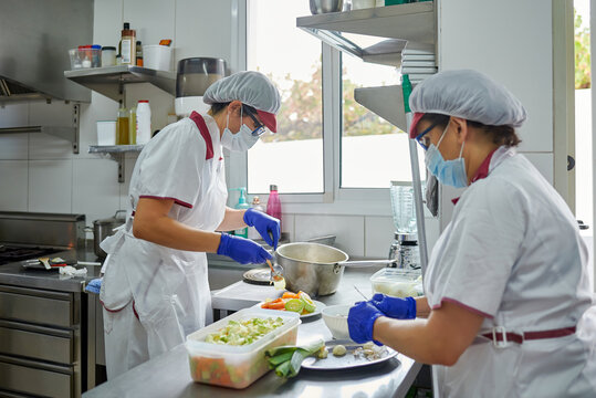 Side view of unrecognizable female cooks in protective masks and gloves preparing meal for patients while working in hospital kitchen during coronavirus pandemic