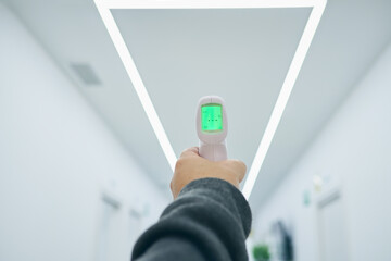 Crop anonymous male standing in white corridor of modern hospital with infrared thermometer for measuring temperature during coronavirus epidemic