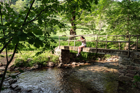 Calm Female Sitting On Old Bridge Over River In Woods And Enjoying Nature In Summer