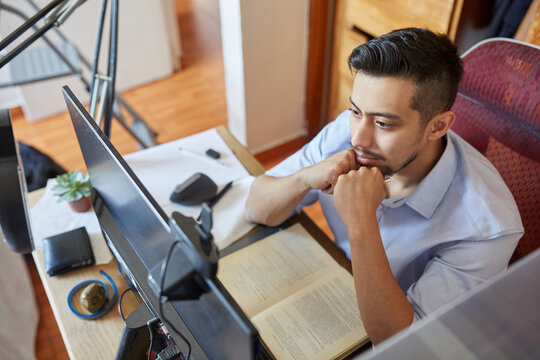 High angle side view of young male student sitting at table and working on computer monitor while preparing for exam in university