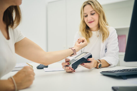Crop person making payment with smartphone in POS terminal in hands of smiling cosmetologist in beauty center