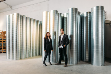 Smiling businesspeople in formal wear standing in contemporary industrial warehouse with metal barrels and looking at camera