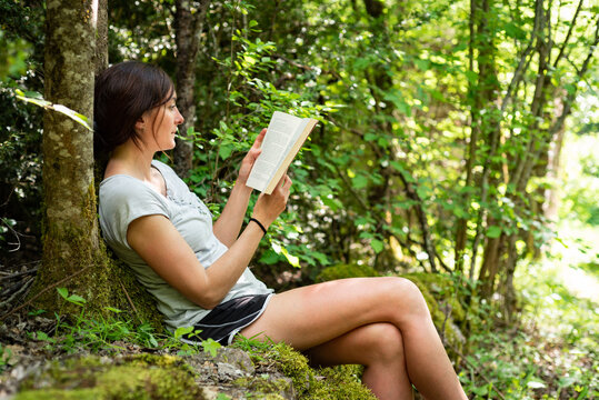 Side view of focused female sitting on mossy stones in woods and enjoying interesting story in book while spending time in nature