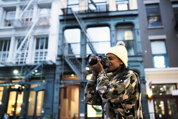 Low angle of African American female photographer in warm hat and stylish sweater taking picture of modern photo camera in New York city