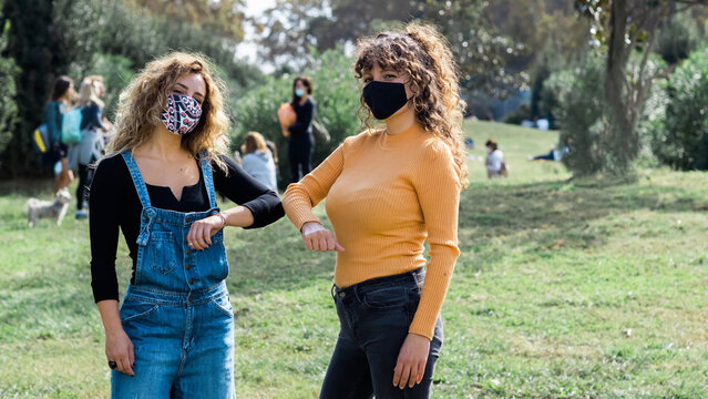 Female friends wearing protective masks bumping elbows while greeting each other during coronavirus epidemic