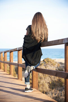 Back view of unrecognizable calm brunette in cozy sweater with long hair standing at on wooden promenade the beach