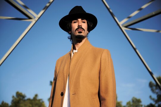 From below of confident young ethnic male in stylish coat and hat looking at camera while standing against high metal electric tower and blue sky