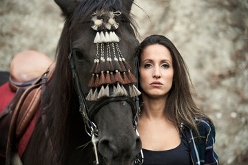 Peaceful female equestrian hugging chestnut horse in bridle while standing in nature and looking at camera