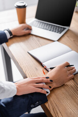 cropped view of secretary touching hand of businessman near notebook and laptop, blurred background