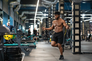 Fototapeta na wymiar Shirtless muscular black guy jumping with skipping rope in gym