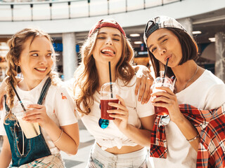 Three young beautiful smiling hipster female in trendy summer clothes. Sexy carefree women in shopping center. Models holding and drinking fresh cocktail smoothie drink in plastic cup with straw