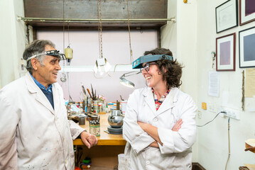 Positive middle aged male and female colleagues in white work coats and professional glasses discussing news and laughing while having break during work in jewelry workshop