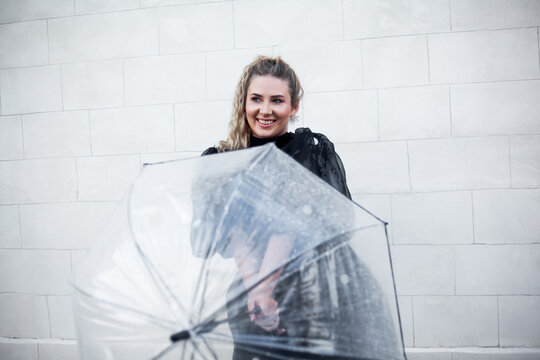 Style woman dressed, black dress walking in the street against the city white wall and hiding under a transparent umbrella from the rain