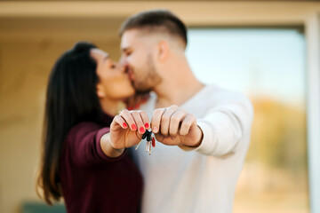 Young couple with keys kissing and showing keys to camera on background of new house