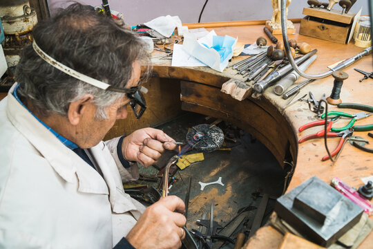 High angle side view of skilled mature male master in glasses using soldering iron while working at workbench with professional instruments in jewelry workshop