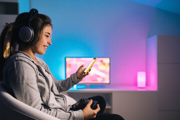 Side view of cheerful woman in headphones and with joystick sitting in armchair at home and browsing cellphone