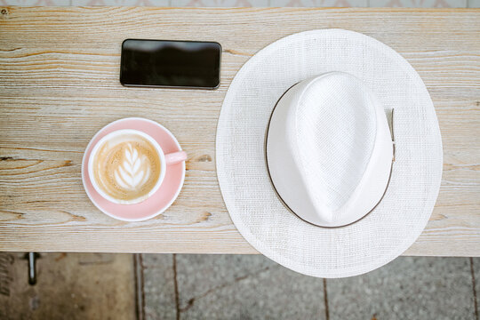 From above of cup of delicious cappuccino with coffee art arranged on wooden bench with mobile phone and white stylish hat