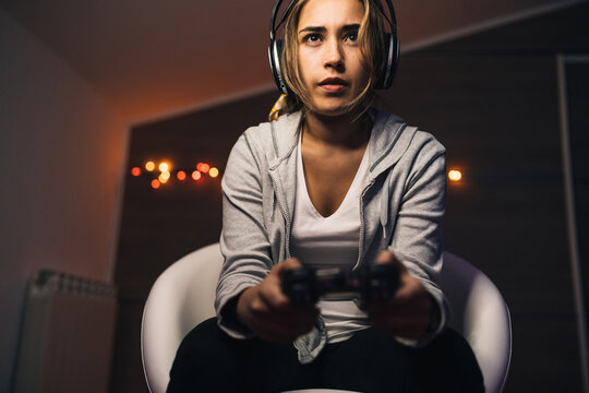 From below concentrated female in headphones and with console sitting in armchair and playing video game while entertaining in evening at home