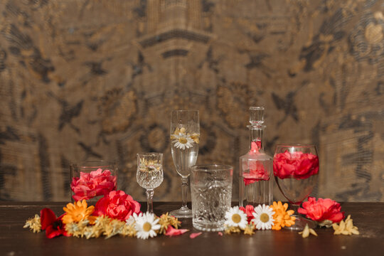 Beautiful stylish arrangement with various types of crystal glasses and bottle placed among assorted fresh flower buds on table against blurred ornamental background