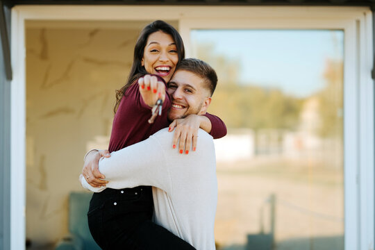 Side view of cheerful couple with keys hugging and looking at camera on background of new house