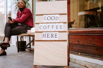 Wooden board with inscription Good Coffee Here placed near coffee shop on background of Stylish African American male sitting on bench near coffee shop browsing on mobile phone on the street