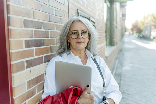 Confident middle aged female entrepreneur in smart casual style standing with laptop along street and looking at camera